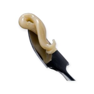 Golden-beige cannabis concentrate on a metallic knife, set against a black background with a retro sunset logo in the corner.